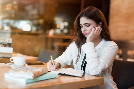 穿着白色时髦夹克的年轻漂亮女人在办公室外工作，在咖啡厅休息时在策划书上写字，看起来很累的现代女商人在多任务处理中