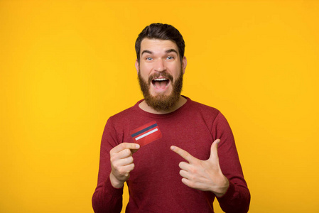 快乐的大胡子年轻人指着一张黄色背景的信用卡。