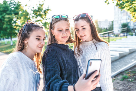 三个十几岁的女孩，在暑期城市的青少年在大学毕业后休息，手中拿着智能手机，读短信写视频通话，在社交网络上在线应用。快乐是微笑的。