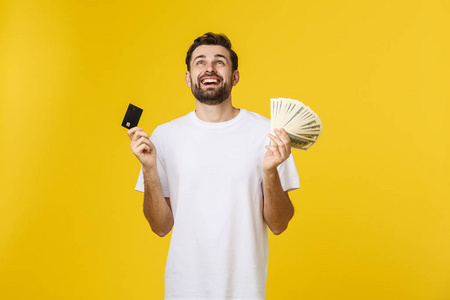 年轻英俊快乐面带微笑的年轻男子，手里拿着银行卡和现金，被隔离在黄色背景上。