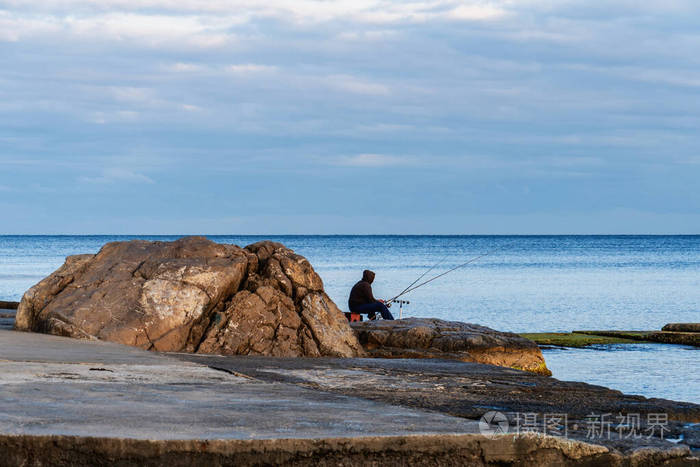 一个孤独的渔夫拿着钓竿坐在海边的岩石上