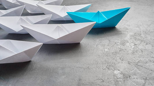 企业领导理念，纸船，关键意见领袖，影响力概念。一艘蓝色的纸船作为领队，在灰色的混凝土背景上朝白色船只的方向前进，