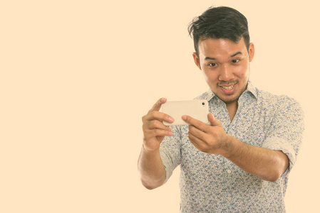 摄影棚拍摄年轻快乐的亚洲男人一边用手机拍照一边微笑