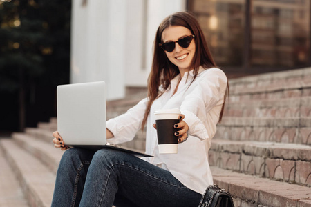 一个穿着牛仔裤和白衬衫的时尚女孩的肖像，拿着笔记本电脑和热咖啡，自由职业者，商务女士，女性权力概念
