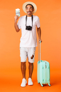 全长垂直拍摄兴奋的非洲裔美国人梦想假期，抬头愉快，想象旅行，持有护照和机票，行李，橙色背景