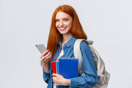 魅力四射的欧洲红发女大学生，背着背包和笔记本，下课回家，用智能手机给朋友发短信，微笑相机