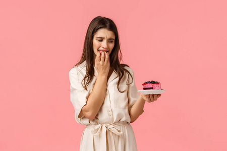 无法抵挡诱惑。少妇在节食，试着吃健康的食物，想吃一口美味的蓝莓蛋糕，咬着指甲盯着盘子，渴望着站在粉红色的背景上