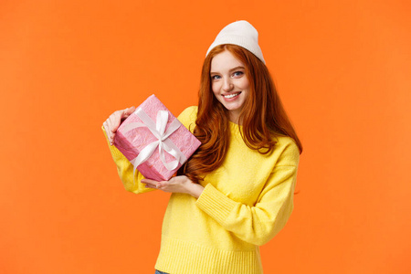 可爱而浪漫可爱的红发女人为情人节准备了礼物，用粉红色的纸包着礼物，展示了盒子里的朋友和欢快的微笑，站在橙色背景上戴着冬天的帽子