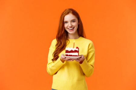 女朋友搞了个惊喜派对，带上一块蛋糕和一支蜡烛庆祝伴侣生日，唱着b日快乐和可爱的微笑，站在黄色毛衣的橙色背景上