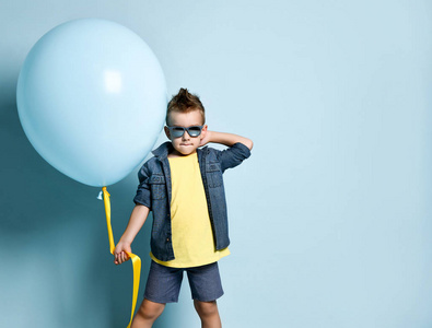 一个金发男孩穿着时髦的休闲服装，穿着运动鞋，戴着墨镜，站在蓝色气球旁边看着镜头