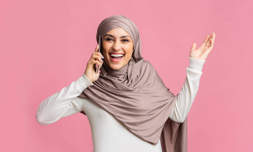 惊讶的伊斯兰女孩戴着头巾在粉红色的背景上打电话