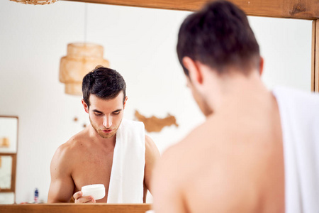 肩上扛着毛巾，手里拿着一罐奶油的运动型年轻人站在浴室镜子前