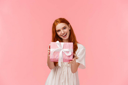 给你。可爱而温柔，女性化的红发女孩送给你她的礼物，拿着包装好的盒子，祝贺生日，情人节快乐，微笑着站在粉红色的背景下
