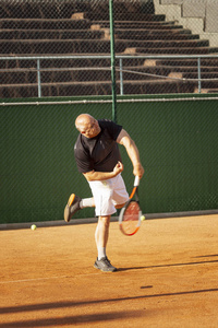 阳光明媚的日子，一个秃顶的男人在球场上打网球。健康和活动。