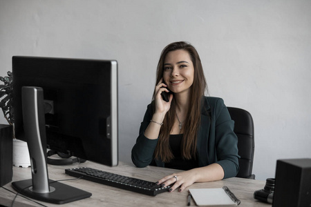 白色办公室里对着电脑屏幕打电话的年轻女商人的画像。女企业家坐在桌子。电话工作电话。
