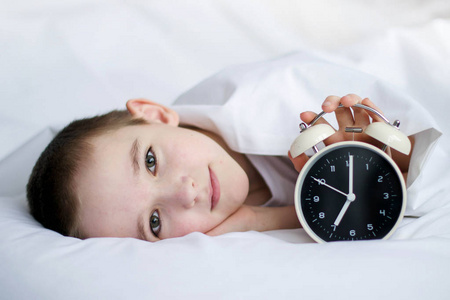男孩躺在一张白色的床上，手里拿着闹钟。这个