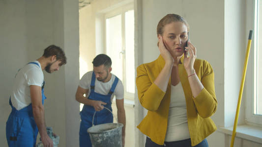 Woman talks on phone, builders in uniform work 