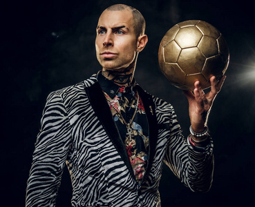 大胆纹身的男模在黑暗的工作室里摆姿势拍照，一边拿着一个金色的足球，一边把视线移开