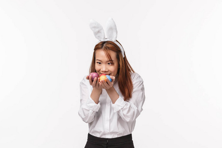 复活节假期，庆祝概念。有趣的亚洲女人在兔子耳朵里，抱着贪婪和惊讶的彩蛋，仿佛在告诉我珍贵的，站着娱乐的白色背景