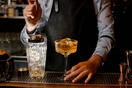 穿着黑色围裙的酒吧招待小心地在玻璃杯上洒上棕色酒精鸡尾酒。