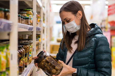 戴医用口罩的年轻女子在超市挑选罐头食品