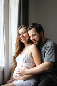 快乐的白人丈夫在窗边拥抱怀孕的妻子。