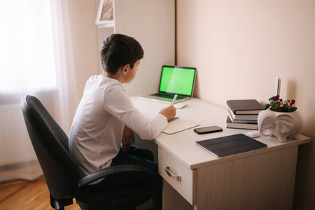 小学生在他房间的书桌旁学习。男孩用笔记本电脑，在笔记本上写字。桌子上的书和写字板。在qurantine期间回家学习。绿色屏幕