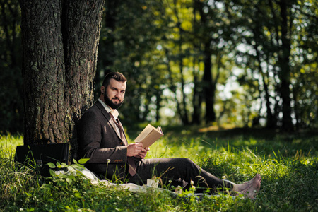 年轻迷人的大胡子商人坐在树下的绿草上，在公园里休息。读书，喝咖啡。放松，厌倦工作，午休。