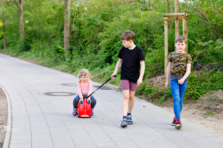 三个孩子，一个蹒跚学步的小女孩和两个男孩在公园里散步。孩子们，可爱的兄弟姐妹们一起玩耍，用玩具汽车行李箱装着轮子