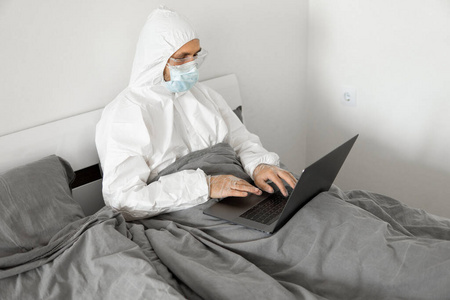 由于冠状病毒流行，一名身穿白色防护服戴着医用面罩的男子正拿着笔记本电脑在家里工作。大流行期间的远程工作。在COVID19隔离