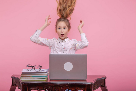 美丽的年轻女孩摇着长长的金发，坐在电脑前欣喜若狂。在粉红色的背景上。