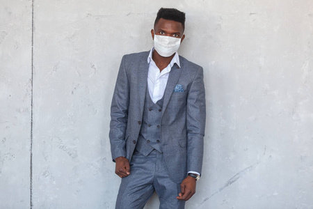 科维德19。一位自信的年轻非洲男子穿着灰色西装，靠在水泥墙上，戴着一个防护性的医用面罩，以防感染冠状病毒和流感大流行