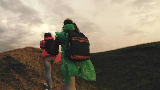 强壮健康的女孩旅行者们穿着五颜六色的雨衣，爬山，牵手，互相帮助。免费女游客背着背包登上山顶，为胜利而欢欣雀跃