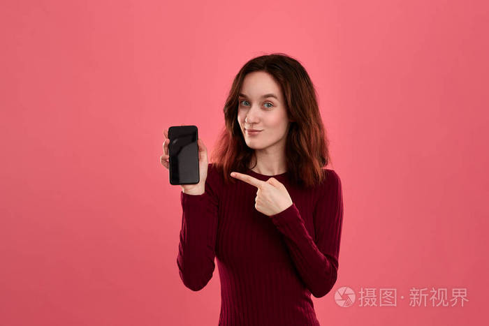 站在深粉色背景上看着相机兴奋的棕发女孩用手指指着手机屏幕