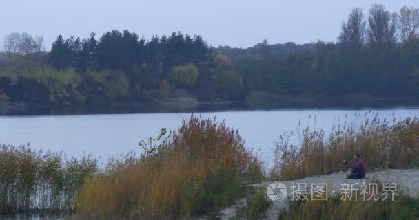 男读一本书坐在附近河看着水桑迪银行的湖景观树木剪影黄昏绿色和黄色草藤生长光滑水秋