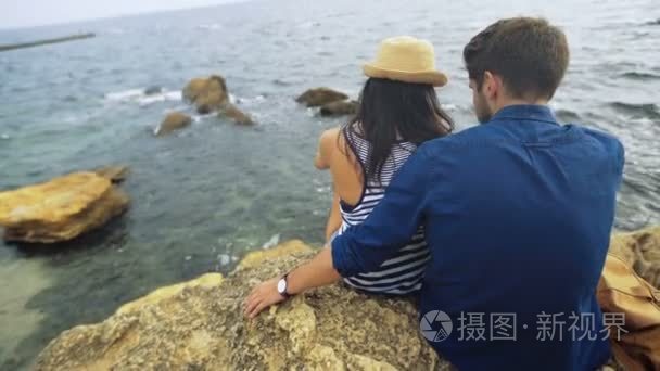 两个年轻快乐旅游坐在石头上看着平静的海面。抱着他的女人的男人