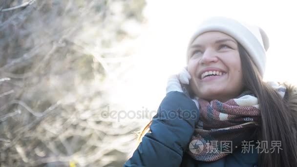 使用智能手机在严寒的冬天室外的美女。跟某人通过手机在阳光明媚的早晨我站在公园的小女孩微笑着。自然背景视频