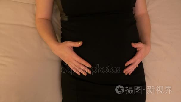 特写镜头的年轻怀孕的准妈妈，穿件黑衣服，躺在床上按摩她的肚子