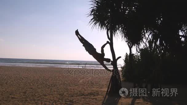 年轻的家伙演示人类旗子在海滩。健壮的男人做体操元素在异国情调的大洋彼岸的棕榈树上。男运动员在训练期间执行力量练习。外面的培训