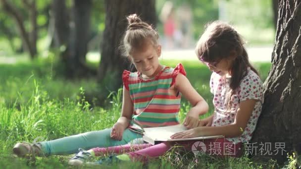 两个小女孩在树下享受阳光明媚的日子和看书。姐姐给她妹妹读一本书
