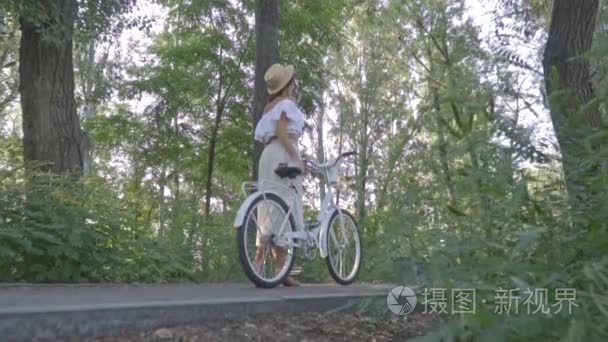 相机从后面突袭上纤细的女孩，在一条白色的裙子，上衣和一顶草帽，站在一个公园与城市自行车，找人的人之中，回视图中。慢动作视频