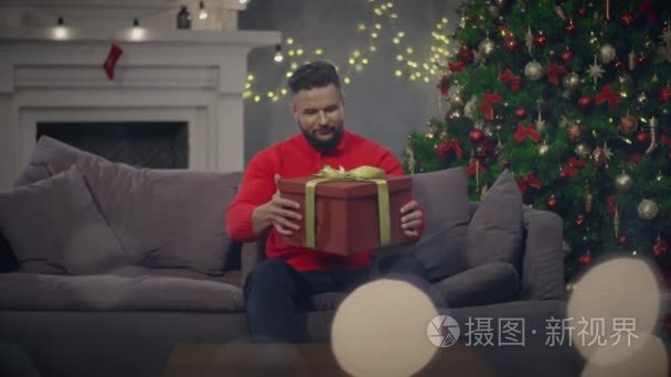 年轻人与圣诞节礼物在手打开它坐在沙发上