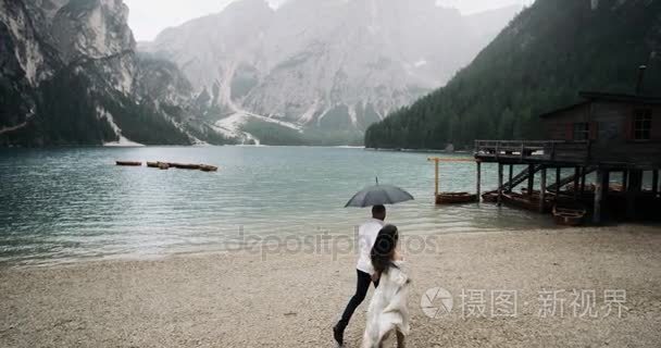 梦幻般的景观与大湖和山在雨天与夫妇运行在伞下的木房子。慢动作4k. 红色史诗。Braies