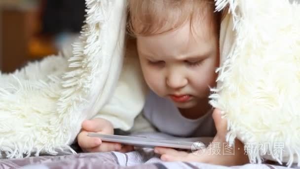 孩子看起来卡通和播放下载的应用程序在一个智能手机特写。一个可爱有趣的小女孩躺在床下的毯子  看着白色的电话屏幕