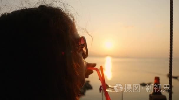 年轻漂亮的女人在太阳镜享受热带龙舌兰酒在海边  在她的暑假美丽的日落装饰花的酒精鸡尾酒。慢动作。1920x1080