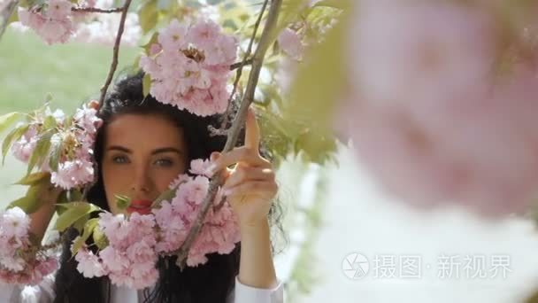 美丽的女人看在相机通过粉红色的樱花花