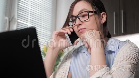 工作中的自由职业者。 漂亮的年轻女人戴着眼镜，在厨房里打电话，看着笔记本电脑，同意