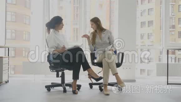 办公室工作人员，两个女人坐在椅子上说话，其中一个女人讲一个有趣的故事，另一个人笑