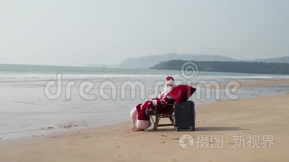 沙滩上的圣诞老人坐在手提箱旁休息。 海边的圣诞节