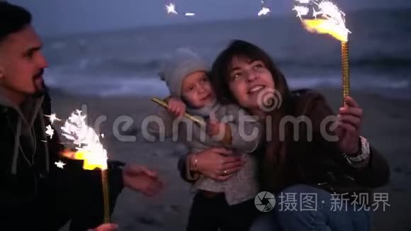 小女孩在父亲的怀里`看着她的母亲，父亲在黄昏的海滩上挥舞着火花。 年轻快乐